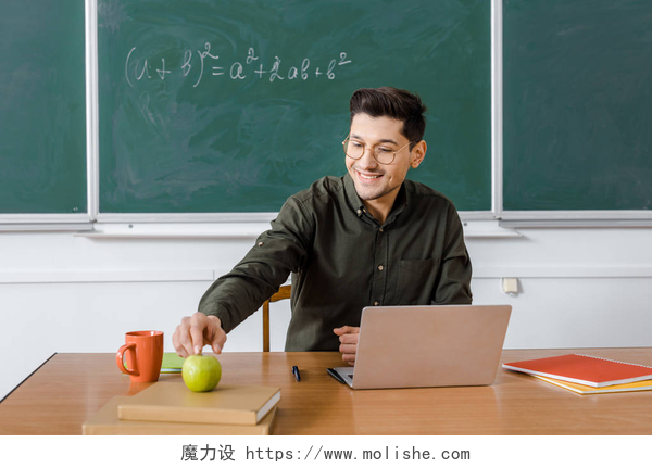 微笑的男老师戴着眼镜坐在讲台前面微笑的男老师戴着眼镜坐在电脑桌前, 伸手去拿教室里的苹果